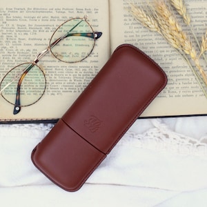 May include: A brown leather glasses case with a gold embossed logo on a white crocheted surface with a book and dried wheat stalks.