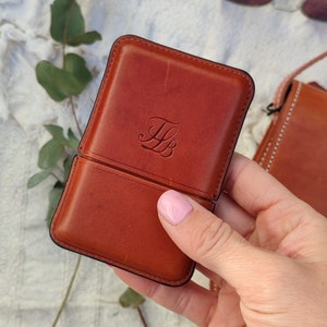May include: A brown leather case with a black border, featuring the initials "FB" embossed on the front. The case is open, revealing the interior. A pair of sunglasses is visible in the background.