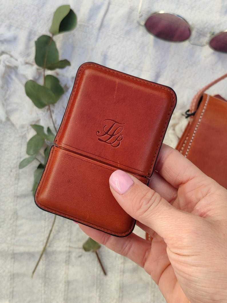 May include: A brown leather case with a black border, featuring the initials "FB" embossed on the front. The case is open, revealing the interior. A pair of sunglasses is visible in the background.