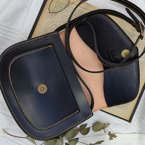 May include: A navy blue leather crossbody bag with a rounded flap and a contrasting light pink interior. The bag features gold stitching and a magnetic clasp. The strap is adjustable. The bag is open, revealing the interior.