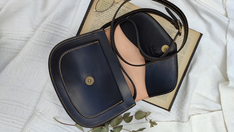 May include: A navy blue leather crossbody bag with a rounded flap and a contrasting light pink interior. The bag features gold stitching and a magnetic clasp. The strap is adjustable. The bag is open, revealing the interior.