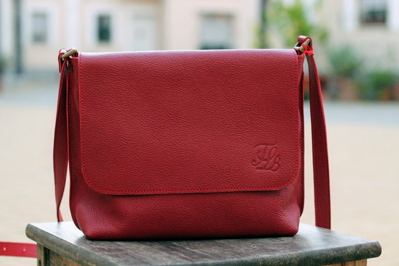 May include: A red leather messenger bag with a flap closure and a gold metal clasp. The bag has a small embossed letter "B" on the front.