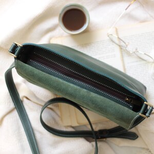 May include: A green leather crossbody bag with a zipper closure and a suede lining. The bag has a long adjustable strap.