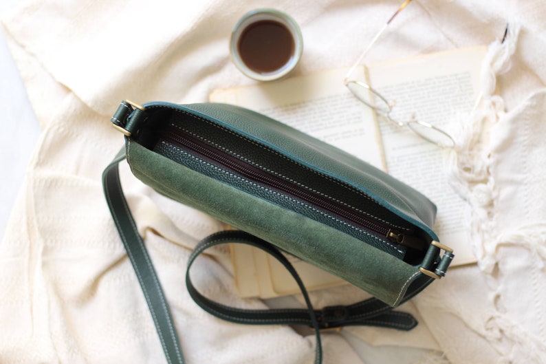 May include: A green leather crossbody bag with a zipper closure and a suede lining. The bag has a long adjustable strap.