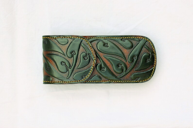 May include: A green leather case with a brown embossed pattern of leaves and swirls. The case has a yellow stitch around the edges.