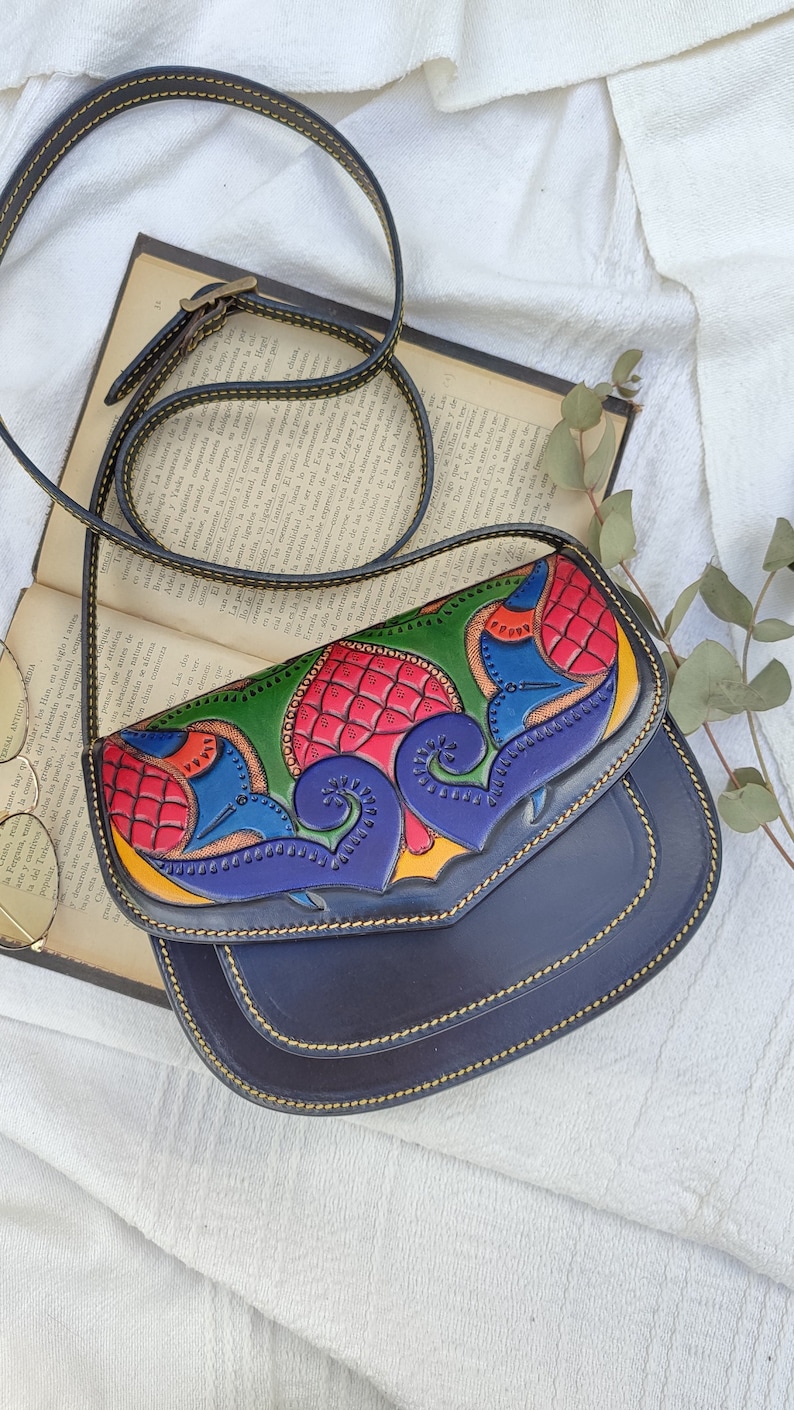 Puede incluir: Un pequeño bolso bandolera de cuero azul oscuro con cierre de solapa. La solapa presenta un diseño colorido e intrincado de flores y remolinos en tonos rojos, amarillos, verdes y azules.