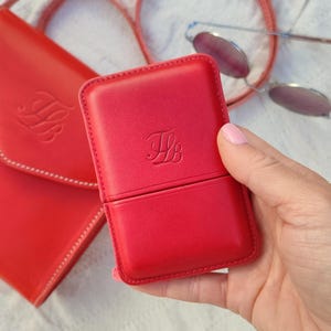 May include: A red leather case with a flap, held in a hand. The case has a debossed monogram and is next to a matching red purse and a pair of round sunglasses. The case is rectangular and appears to be a glasses case or similar.