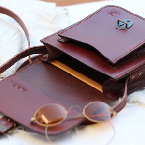 K&ouml;nnte beinhalten: Eine braune Leder-Umh&auml;ngetasche mit Klappenverschluss und einer Metallschnalle. Die Tasche ist offen und hat ein beigefarbenes Futter. Ein Paar runde Sonnenbrillen befindet sich in der Tasche.