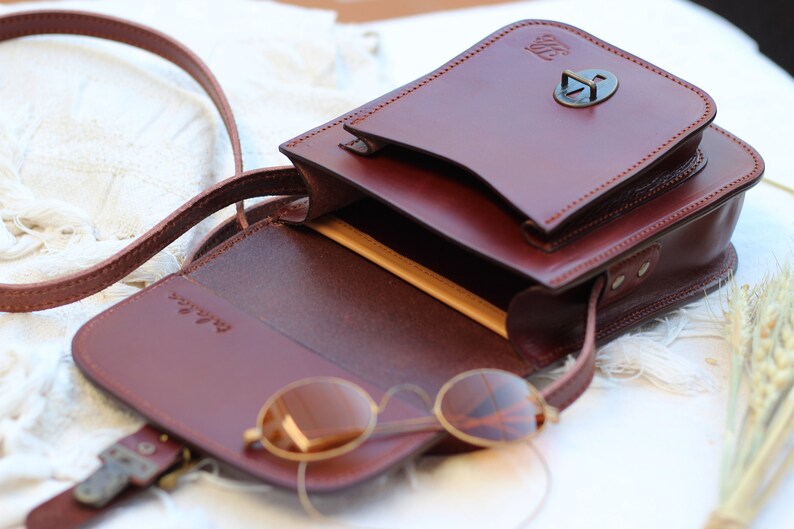 K&ouml;nnte beinhalten: Eine braune Leder-Umh&auml;ngetasche mit Klappenverschluss und einer Metallschnalle. Die Tasche ist offen und hat ein beigefarbenes Futter. Ein Paar runde Sonnenbrillen befindet sich in der Tasche.