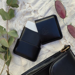 May include: Black leather card holder with an embossed monogram, open to reveal a white card. A pair of sunglasses with pink lenses and a black leather purse are also in the image. The items are arranged on a white, textured surface.