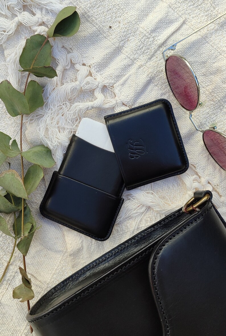 May include: Black leather card holder with an embossed monogram, open to reveal a white card. A pair of sunglasses with pink lenses and a black leather purse are also in the image. The items are arranged on a white, textured surface.