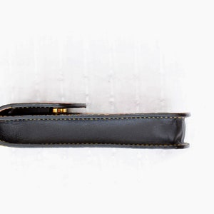 Op de afbeelding: Een zwart leren etui met een gouden sluiting en gele stiksels. Het etui is gesloten en ligt op een wit oppervlak.