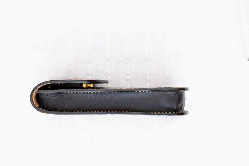 Op de afbeelding: Een zwart leren etui met een gouden sluiting en gele stiksels. Het etui is gesloten en ligt op een wit oppervlak.