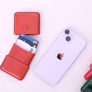 May include: An arrangement of leather wallets in various colors, including red, green, and burgundy, alongside a white smartphone. One red wallet is open, displaying credit cards. The wallets have a minimalist design, and the phone features the Apple logo.