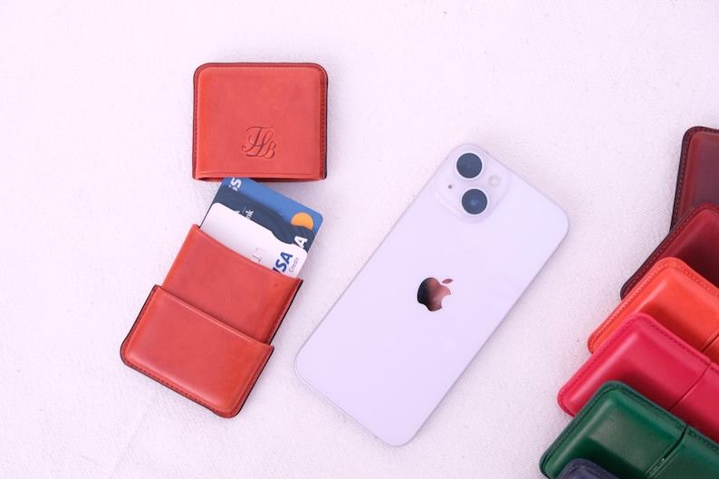 May include: An arrangement of leather wallets in various colors, including red, green, and burgundy, alongside a white smartphone. One red wallet is open, displaying credit cards. The wallets have a minimalist design, and the phone features the Apple logo.