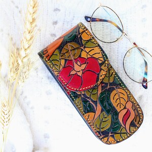 Op de afbeelding: Een leren brilletje met een bloemenpatroon en een rood bloemenontwerp. De etui is gesloten en ligt op een wit oppervlak naast een bril met een gouden montuur.