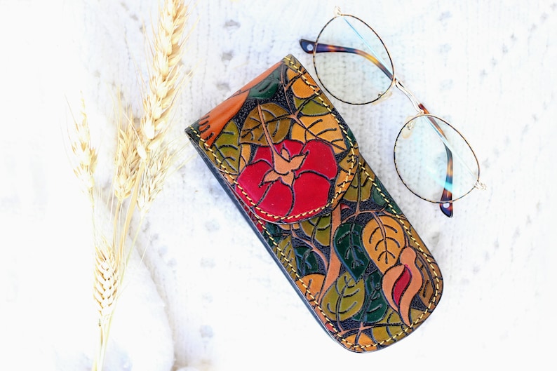 Op de afbeelding: Een leren brilletje met een bloemenpatroon en een rood bloemenontwerp. De etui is gesloten en ligt op een wit oppervlak naast een bril met een gouden montuur.