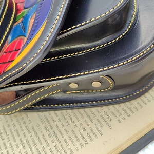 May include: Black leather crossbody bag with a colorful, hand-painted design. The bag features a rounded shape, a long strap, and yellow stitching. The flap has a vibrant, abstract design with red, blue, and yellow accents.