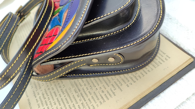May include: Black leather crossbody bag with a colorful, hand-painted design. The bag features a rounded shape, a long strap, and yellow stitching. The flap has a vibrant, abstract design with red, blue, and yellow accents.