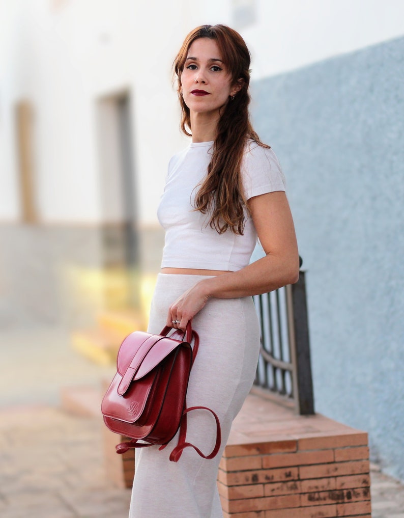 May include: A woman wearing a white crop top and a beige skirt is holding a red leather backpack. The backpack has a flap closure and a small pocket on the front.