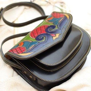 May include: Black leather crossbody bag with a colorful, hand-painted flap. The flap features a detailed design with red, green, blue, and yellow accents. The bag has a long, adjustable strap and gold stitching.
