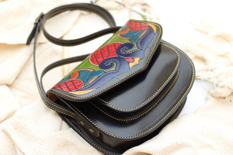 May include: Black leather crossbody bag with a colorful, hand-painted flap. The flap features a detailed design with red, green, blue, and yellow accents. The bag has a long, adjustable strap and gold stitching.