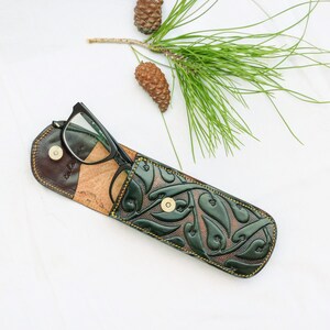 May include: A green leather glasses case with a floral pattern and a brown leather flap. The case is open and a pair of black glasses is inside.