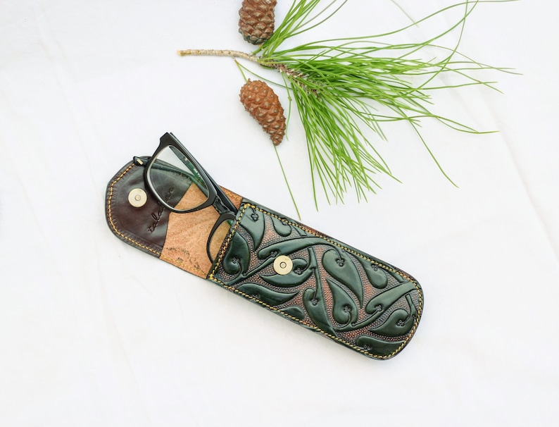May include: A green leather glasses case with a floral pattern and a brown leather flap. The case is open and a pair of black glasses is inside.