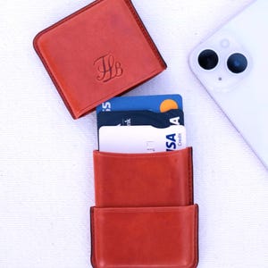 May include: A burnt orange leather card holder with a matching top cover, displaying the initials "TB". The card holder is open, revealing several credit cards. A white smartphone is in the background.