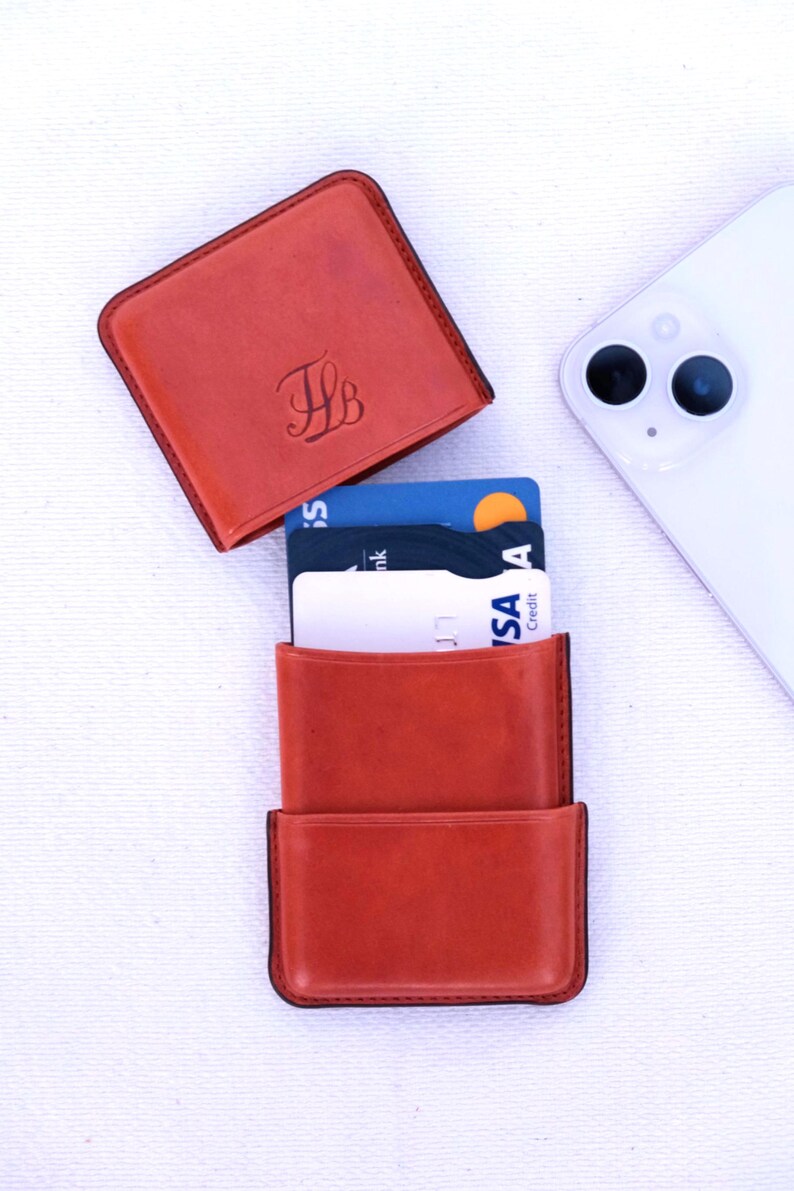 May include: A burnt orange leather card holder with a matching top cover, displaying the initials "TB". The card holder is open, revealing several credit cards. A white smartphone is in the background.