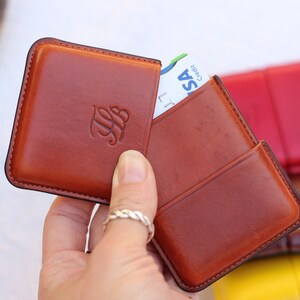 May include: A brown leather cardholder with a raised monogram, holding a credit card. The cardholder is open, revealing the card. Other cardholders in red, brown, and yellow are in the background. The cardholder is held by a person.