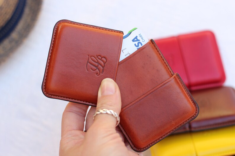 May include: A brown leather cardholder with a raised monogram, holding a credit card. The cardholder is open, revealing the card. Other cardholders in red, brown, and yellow are in the background. The cardholder is held by a person.
