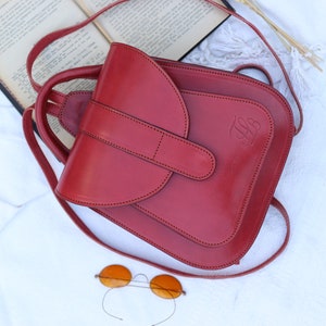 May include: A red leather backpack with a flap closure and a small pocket on the front. The backpack has a strap that can be adjusted to fit different heights. The backpack is sitting on a white surface.