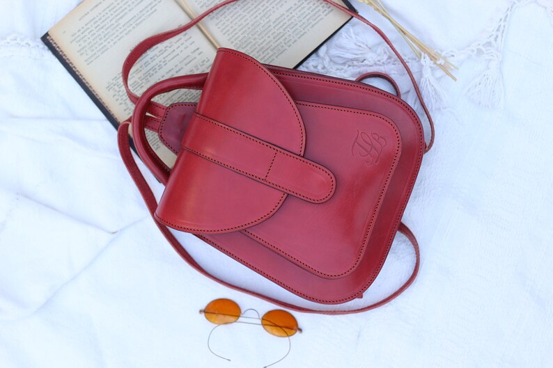 May include: A red leather backpack with a flap closure and a small pocket on the front. The backpack has a strap that can be adjusted to fit different heights. The backpack is sitting on a white surface.