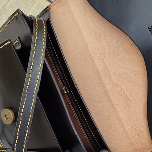 May include: Black leather crossbody bag with a flap closure, featuring yellow stitching and a tan interior. The bag has a long strap and a metal clasp. The interior includes a zippered pocket.
