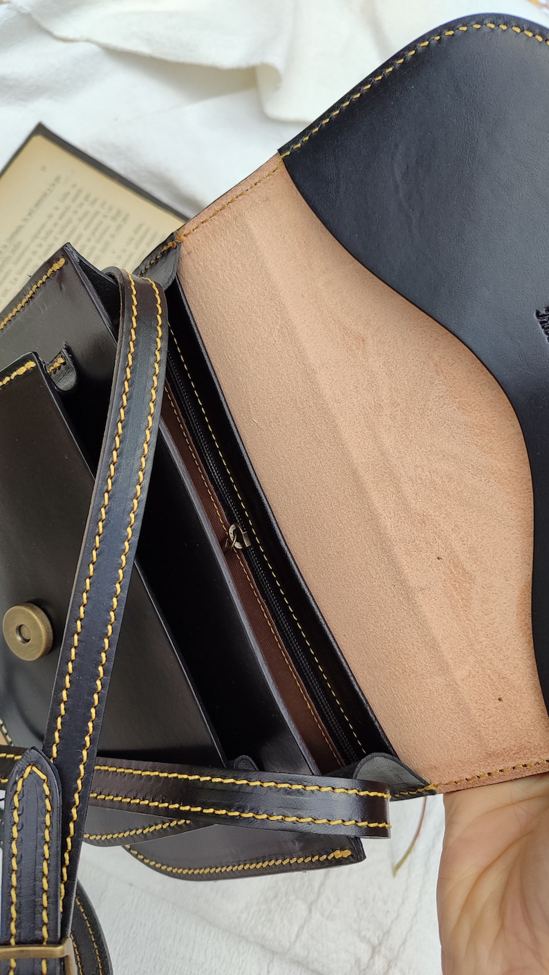 May include: Black leather crossbody bag with a flap closure, featuring yellow stitching and a tan interior. The bag has a long strap and a metal clasp. The interior includes a zippered pocket.