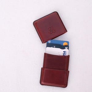 May include: A burgundy leather card holder with a matching square top. The top has an embossed design. The card holder is open, revealing several credit cards. The background is a plain white surface.