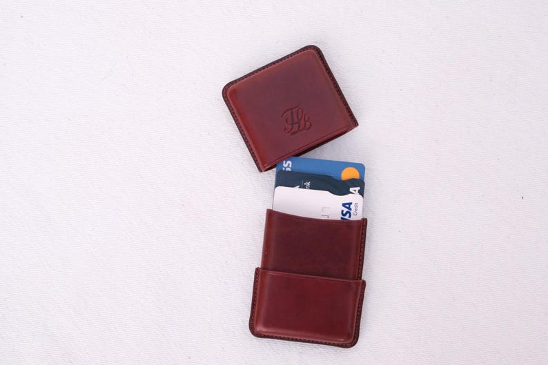 May include: A burgundy leather card holder with a matching square top. The top has an embossed design. The card holder is open, revealing several credit cards. The background is a plain white surface.