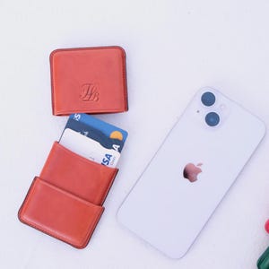 May include: An arrangement of leather cardholders and a smartphone on a white surface. The cardholders are in shades of orange and red, with one open to display credit cards. The smartphone is white with a silver Apple logo.