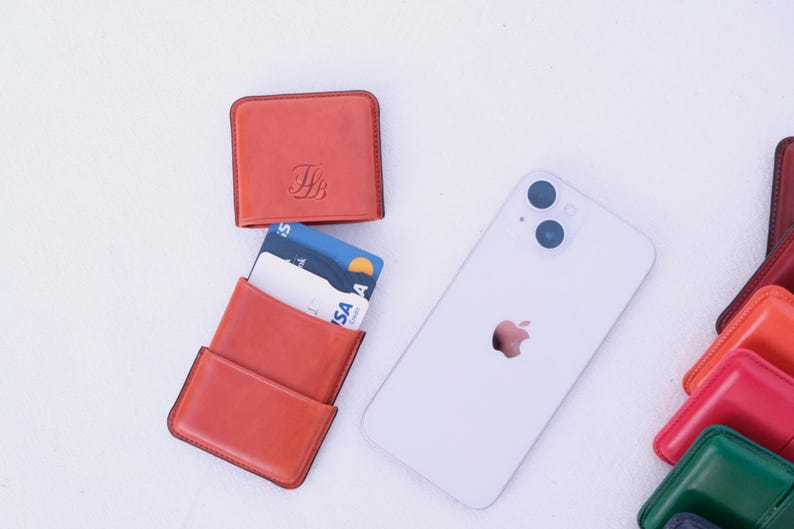 May include: An arrangement of leather cardholders and a smartphone on a white surface. The cardholders are in shades of orange and red, with one open to display credit cards. The smartphone is white with a silver Apple logo.