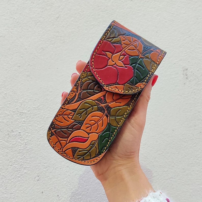 Op de afbeelding: Een bruine leren potlood etui met een bloemenpatroon met rode, oranje en groene bladeren. De etui heeft een klep sluiting.