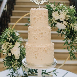 May include: A three-tiered wedding cake with pearl-like decorations and a "Just Married" cake topper. The cake sits on a white pedestal, surrounded by floral arrangements and a gold hoop. A cake knife rests on the table.