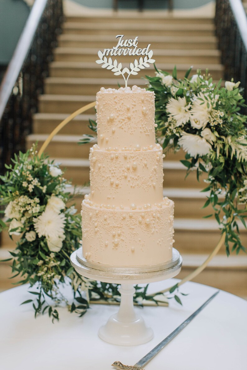 May include: A three-tiered wedding cake with pearl-like decorations and a "Just Married" cake topper. The cake sits on a white pedestal, surrounded by floral arrangements and a gold hoop. A cake knife rests on the table.
