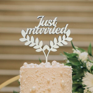 May include: A wooden cake topper with the words "Just Married" in a cursive font, framed by a laurel wreath design. The topper is placed on a light-colored cake decorated with pearls and sprinkles. The background is blurred.
