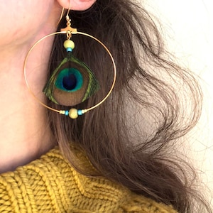 May include: Gold hoop earrings with a peacock feather and small beads in shades of green, blue, and yellow. The earrings are suspended from a gold hook. The person is wearing a yellow sweater.