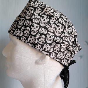 May include: A black and white skull print surgical scrub cap. The cap is tied in the back with a black ribbon. The skull pattern is repeated across the fabric.