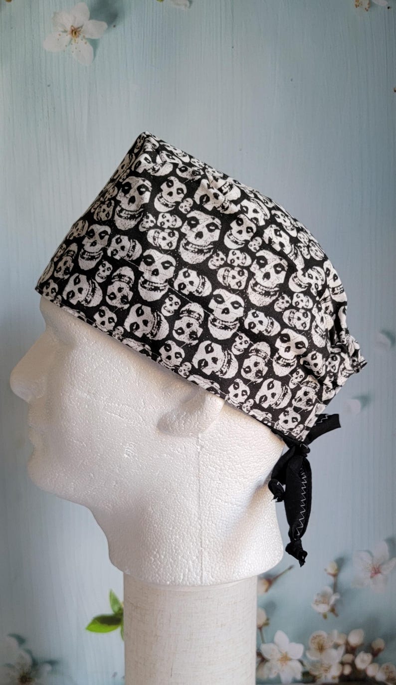 May include: A black and white skull print surgical scrub cap. The cap is tied in the back with a black ribbon. The skull pattern is repeated across the fabric.
