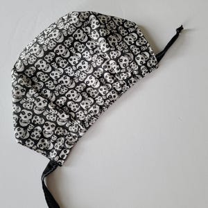 May include: A black and white skull print surgical scrub cap with a black tie. The cap is made of fabric and features a repeating pattern of white skulls on a black background. The tie is made of black fabric.