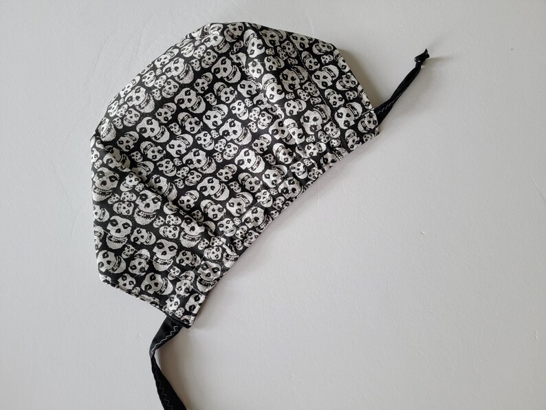 May include: A black and white skull print surgical scrub cap with a black tie. The cap is made of fabric and features a repeating pattern of white skulls on a black background. The tie is made of black fabric.