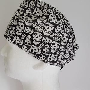 May include: A black and white skull print scrub cap. The cap is made of fabric and is designed to cover the head. The skull pattern is repeated across the entire surface of the cap. The cap is shown on a white mannequin head.